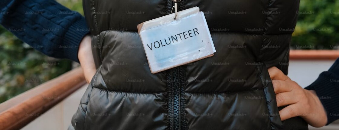 Eine Frau hat eine schwarze Steppweste an und trägt ein Schild mit der Aufschrift "Volunteer".