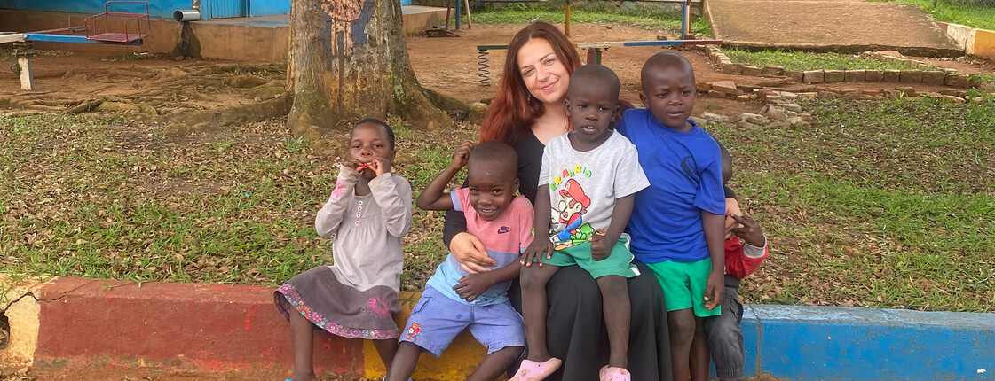 Person sitzt mit mehreren Kindern auf einer bunten Kante im Hof des Nsambya Babies Home.