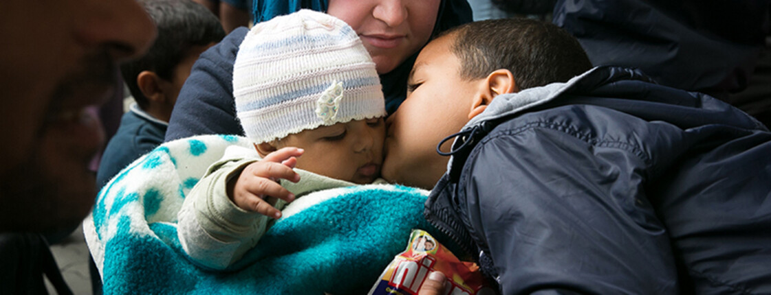 Eine junge Mutter hält ihr kleines Mädchen in der Hand. Der kleine Bruder gibt dem Mädchen ein Bussi auf die Backe. Die Mutter hat den Kopf in Richtung Kinder gesenkt und sieht entspannt zu ihnen.
