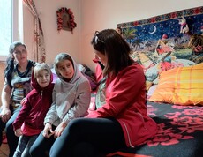Zwei Mädchen und zwei Frauen sitzen auf einer Bettkante. Im Hintergrund ist ein bunter Wandteppich.