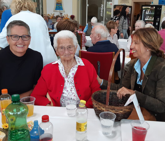 Eine Seniorin in roter Jacke sitzt mit zwei Frauen am Tisch, umgeben von Getränken und herbstlicher Deko.