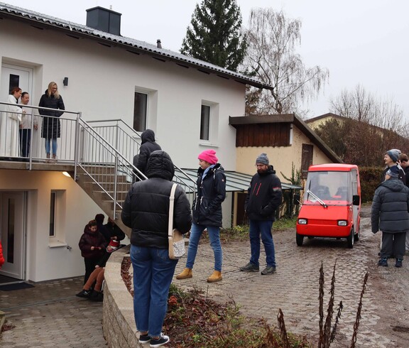 Neues Haus von außen, mit mehreren Menschen und einem roten Auto.