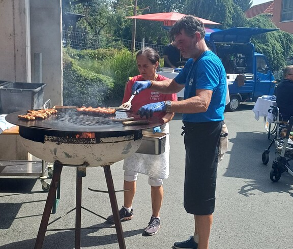 Auf einem großen Rundgrill werden Würstel gegrillt, während zwei Mitarbeitende die Speisen für die Gäste des Sommerfestes im Innenhof des Pflegewohnhauses zubereiten.