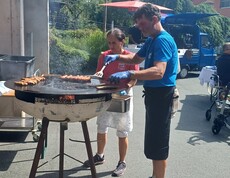 Auf einem großen Rundgrill werden Würstel gegrillt, während zwei Mitarbeitende die Speisen für die Gäste des Sommerfestes im Innenhof des Pflegewohnhauses zubereiten.