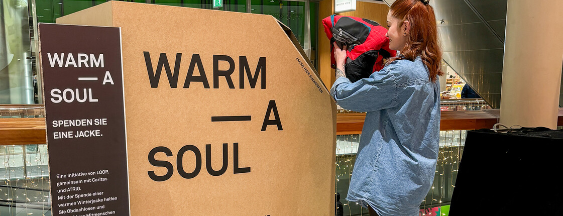 Frau wirft Winterjacke in große Spendenbox mit Text „Warm a Soul“ im Einkaufszentrum ATRIO Villach ein.
