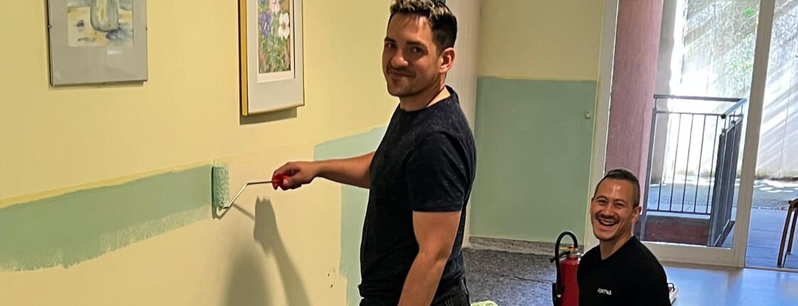 Zwei Männer – einer steht, der andere kniet – lächeln im Gang eines Pflegewohnhauses und blicken direkt in die Kamera. Beide halten eine Farbrolle in der Hand. Sie sind dabei, den unteren Wandbereich in einem freundlichen Mintgrün zu streichen. Die Atmosphäre wirkt fröhlich und engagiert.