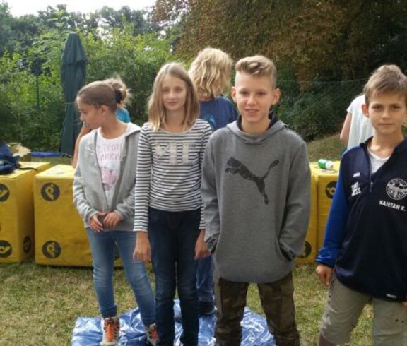 Mehrere Kinder stehen bei einer Gruppenaktivität auf einer Plane im Freien und nehmen gemeinsam an einem Team- und Geschicklichkeitsspiel teil.