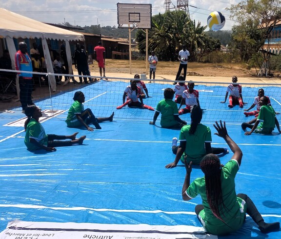 Frauenmannschaften spielen Sitzvolleyball auf einem Spielfeld im Freien, während Zuschauer*innen und Schiedsrichter das Spiel verfolgen.