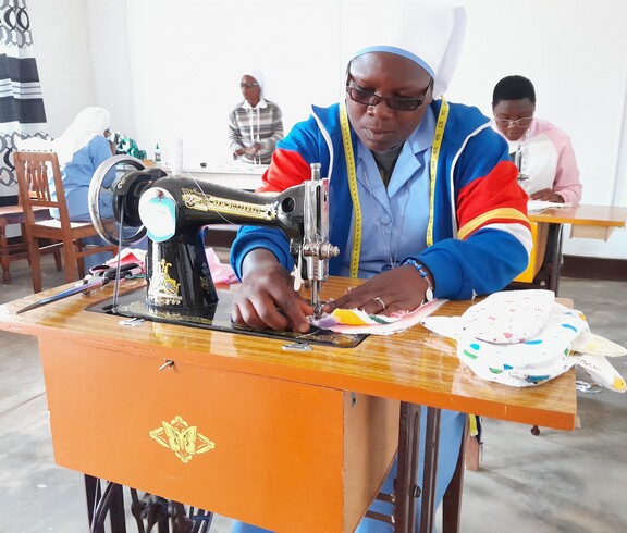 Eine Ordensschwester mit Maßband um den Hals arbeitet konzentriert an einer klassischen Nähmaschine; im Hintergrund sind weitere Frauen beim Nähen zu sehen – Nähatelier des Caritas-Projekts in Burundi