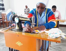 Eine Ordensschwester mit Maßband um den Hals arbeitet konzentriert an einer klassischen Nähmaschine; im Hintergrund sind weitere Frauen beim Nähen zu sehen – Nähatelier des Caritas-Projekts in Burundi