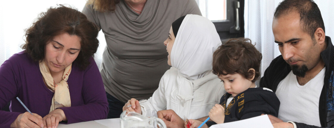 Eine Frau mittleren Alters lernt mit einer Flüchtlingsfamilie. Auf dem Tisch befinden sich Blätter, Bücher und Stifte. Die Frau steht dabei hinter der Familie. Die Familie sitzt am Tisch.