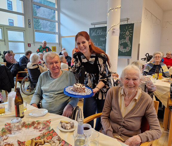 Person präsentiert Kuchen an zwei Gäste, festlich gedeckter Tisch im Pflegewohnhaus