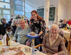 Person präsentiert Kuchen an zwei Gäste, festlich gedeckter Tisch im Pflegewohnhaus