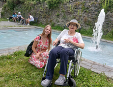 Bewohnerin im Rollstuhl und Schülerin verbringen Zeit miteinander an einem Brunnen im Park.