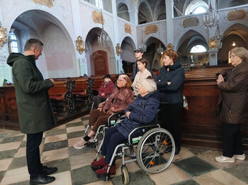 Gruppe älterer Menschen, einige in Rollstühlen, hören dem Pfarrer in einer Kirche zu.