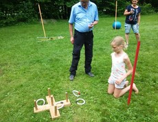 Ein Kind wirft bei einem Geschicklichkeitsspiel Ringe auf Holzstäbe, während weitere Teilnehmende die Aktivität auf einer Wiese begleiten.
