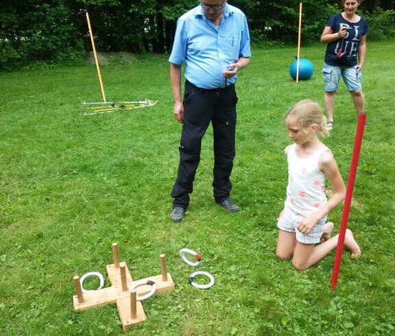 Ein Kind wirft bei einem Geschicklichkeitsspiel Ringe auf Holzstäbe, während weitere Teilnehmende die Aktivität auf einer Wiese begleiten.