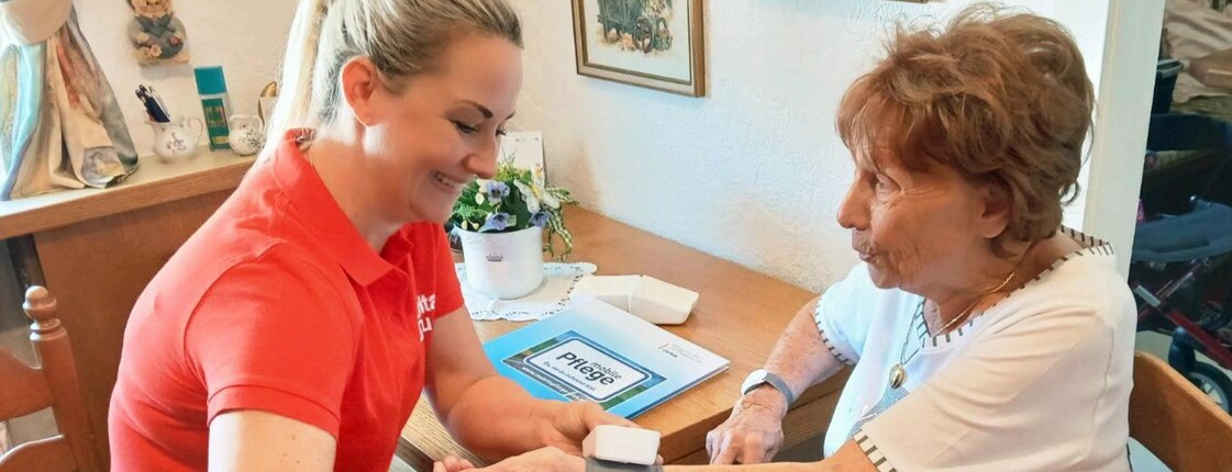 Frau links im Bild im roten T-Sirt misst Blutdruck bei älterer Frau rechts im Bild.Beide sitzend.