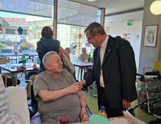 Ein Bewohner sitzt im Aufenthaltsraum des Caritas Pflegewohnhauses, während Weihbischof Johannes Freitag die Person mit einem Handschlag begrüßt und sich im persönlichen Gespräch austauscht.