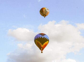 Zwei Heißluftballons schweben vor einem blauen Himmel mit weißen Wolken. Die Aufnahme vermittelt Weite, Ruhe und Leichtigkeit.