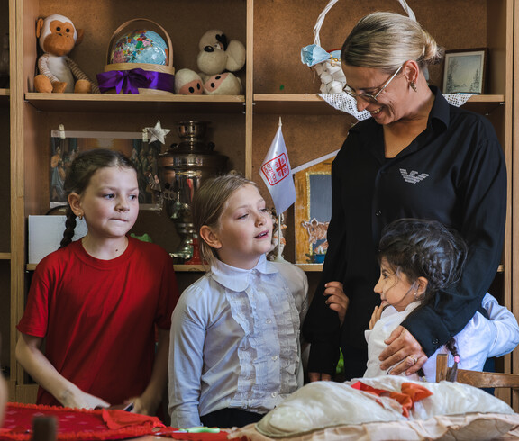 Kinder stehen vor einem Regal mit Spielsachen. Ein Kind umarmt eine Betreuerin, die die Kinder freundlich anlächelt.