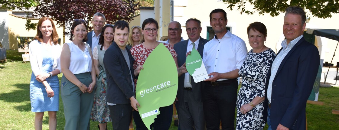 Mehrere Personen stehen schick gekleidet im Garten vor einem Haus und halten ein großes Blatt auf Karton mit der Aufschrift "green care" in die Kamera.