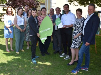 Mehrere Personen stehen schick gekleidet im Garten vor einem Haus und halten ein großes Blatt auf Karton mit der Aufschrift "green care" in die Kamera.