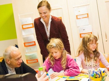 Zwei Erwachsene begleiten Kinder bei einer kreativen Lernaktivität im Klassenraum. Auf dem Tisch liegen Arbeitsmaterialien und Bastelutensilien.