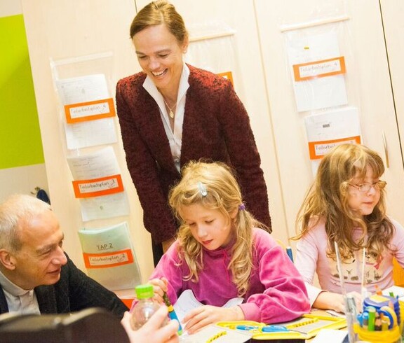 Zwei Erwachsene begleiten Kinder bei einer kreativen Lernaktivität im Klassenraum. Auf dem Tisch liegen Arbeitsmaterialien und Bastelutensilien.