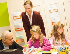 Zwei Erwachsene begleiten Kinder bei einer kreativen Lernaktivität im Klassenraum. Auf dem Tisch liegen Arbeitsmaterialien und Bastelutensilien.