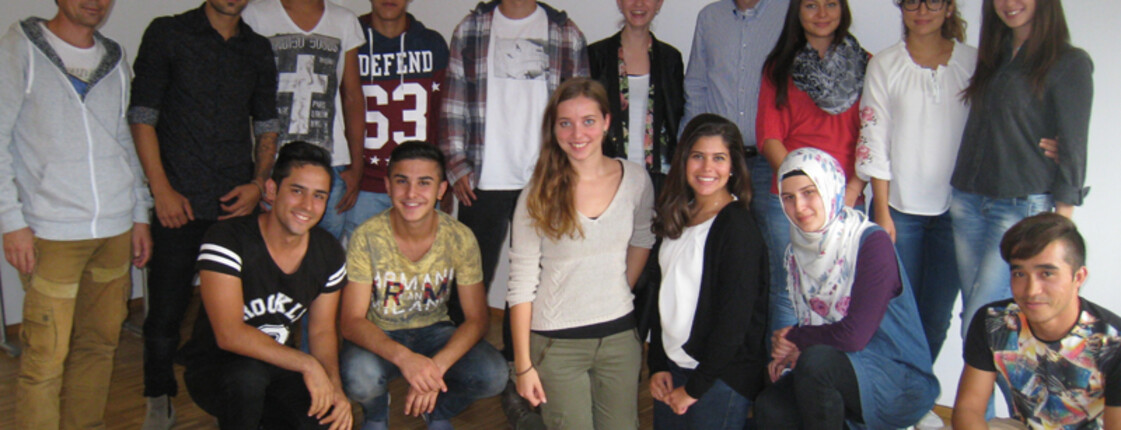 Jugendliche und Erwachsene mit und ohne Migrationshintergrund stehen in einem größeren weißen Raum mit braunem Parkett zu einem Gruppenfoto zusammen. Es sind um die fünfzehn Personen. Alle tragen sie Alltagskleidung.