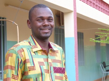 Portrait eines Mannes aus Burkina Faso, der ein buntes Hemd trägt, und vor einem Hauseingang steht.