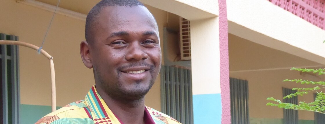 Portrait eines Mannes aus Burkina Faso, der ein buntes Hemd trägt, und vor einem Hauseingang steht.