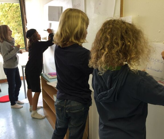 Kinder schreiben gemeinsam Begriffe und Ideen auf große Papierbögen an der Wand und erarbeiten Regeln für das Miteinander im Unterricht.