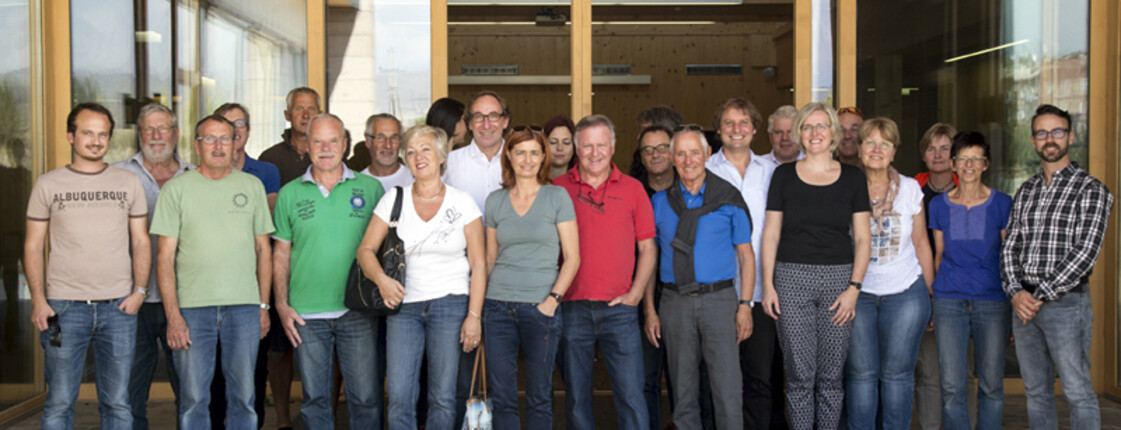 Eine Gruppe vieler Menschen steht vor einem Haus mit großen Glasfenstern. Die meisten tragen eine kurze Kleidung.