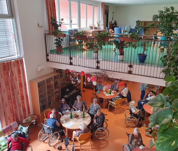 In dem hellen und großzügigen Aufenthaltsbereich eines Pflegewohnhauses sitzen mehrere Bewohnerinnen und Bewohner an Tischen beisammen und genießen das gemeinsame Beisammensein in einer von zahlreichen Pflanzen und herbstlicher Dekoration geprägten Atmosphäre.