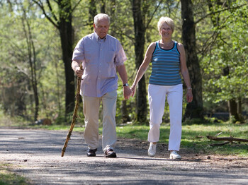 Zwei ältere Personen sind im Wald am Spazieren. Der Mann trägt ein weißes Hemd und eine beige Hose und die Frau eine weiße Hose und ein blaues Top. Der Mann hält zusätzlich einen Stock in der Hand. 