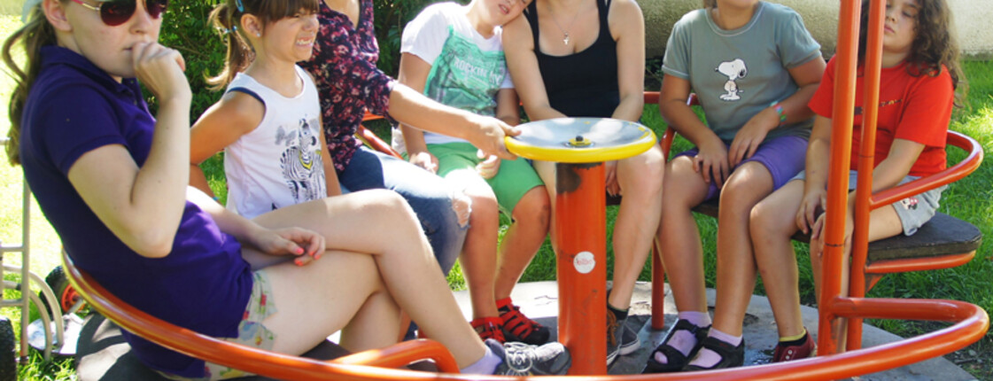 Vier Kinder und drei erwachsene Frauen sitzen in einem orangen Karusell auf einem Spielplatz. Die Kinder, die ca. sechs Jahre alt sind, sind durch das viele Spielen bereits müde und schlaff. Eines lehnt sich an eine Frau an. 