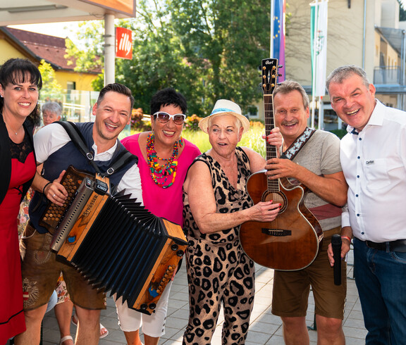 Sechs Personen stehen gemeinsam im Freien. Zwei von ihnen halten Musikinstrumente – eine Person ein Akkordeon, eine andere eine Gitarre. Im Hintergrund hängt ein Banner mit der Aufschrift „20 Jahre“ und „Herzlich Willkommen“. Die Szene wirkt festlich und deutet auf eine Jubiläumsfeier oder ein besonderes Ereignis hin. Von links nach rechts: Claudia Gaube, ein Musiker, Moderatorin Monika Fellner, Jass Gitti, ein Musiker und Josef Url. 