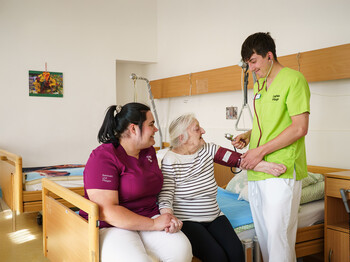 Zwei Pflegekräfte begleiten eine ältere Person beim Blutdruckmessen in einem Pflegezimmer der Caritas.