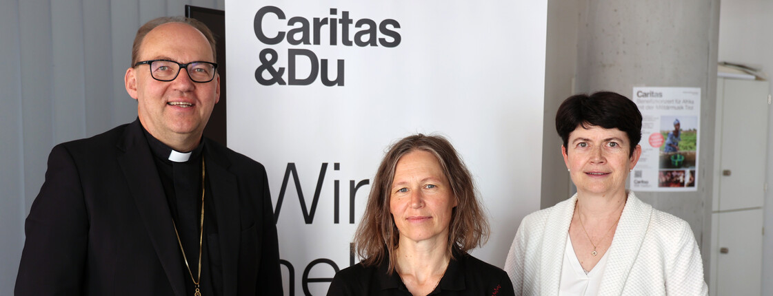 Zwei Frauen und ein Mann stehen nebeneinander vor einem Caritas Roll-Up zu einem Gruppenfoto zusammen.