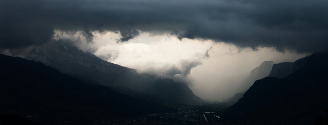 Über einem Tal in den Alpen zieht ein bedrohliches Unwetter mit schwarzen Wolken und Starkregen auf.