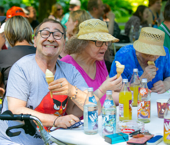 Ein Mann links im Bild mit einer Eistüte in der Hand lacht in die Kamera. Rechts zwei Frauen mit Strohhut essen Eis. Alle drei sitzen an einem Tisch.