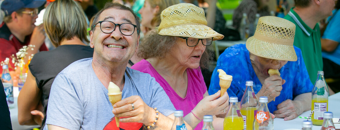 Ein Mann links im Bild mit einer Eistüte in der Hand lacht in die Kamera. Rechts zwei Frauen mit Strohhut essen Eis. Alle drei sitzen an einem Tisch.