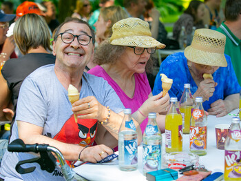 Ein Mann links im Bild mit einer Eistüte in der Hand lacht in die Kamera. Rechts zwei Frauen mit Strohhut essen Eis. Alle drei sitzen an einem Tisch.