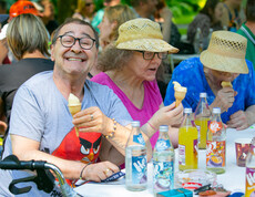 Ein Mann links im Bild mit einer Eistüte in der Hand lacht in die Kamera. Rechts zwei Frauen mit Strohhut essen Eis. Alle drei sitzen an einem Tisch.
