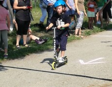 Ein Kind fährt mit Schutzhelm auf einem Roller über die Strecke einer Sportveranstaltung, während Zuschauende am Rand stehen.