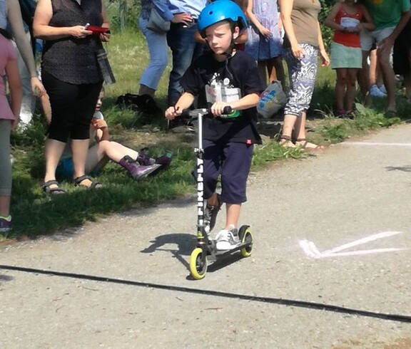 Ein Kind fährt mit Schutzhelm auf einem Roller über die Strecke einer Sportveranstaltung, während Zuschauende am Rand stehen.