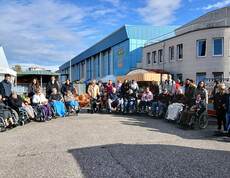 Große Gruppe von Menschen, viele in Rollstühlen, vor einem Gebäudekomplex bei sonnigem Wetter.