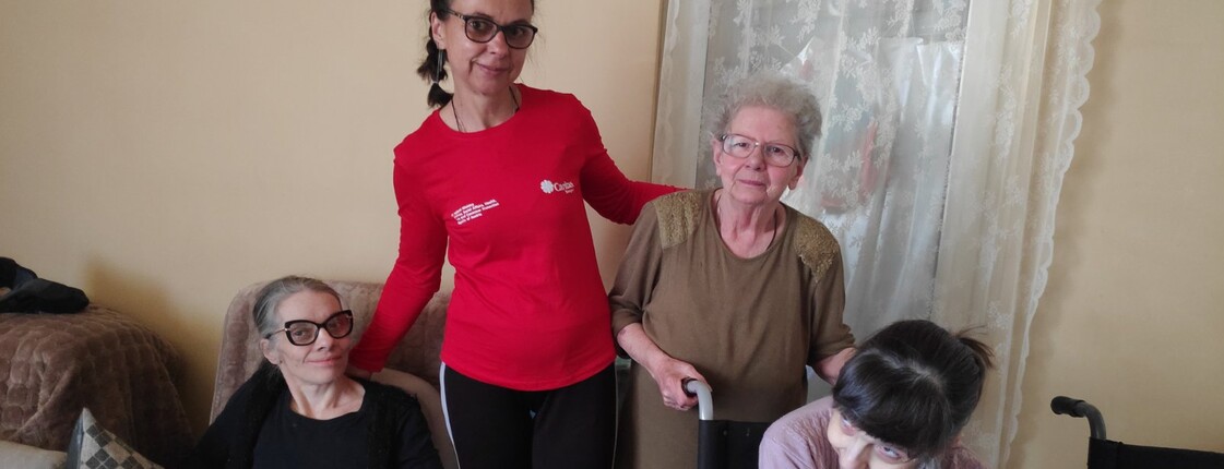 Gruppenfoto: Eine Caritas-Mitarbeiterin in der Mitte mit rotem Caritas-Shirt - daneben eine ältere Dame. Links und rechts zwei jüngere Frauen, die sitzen. Alle lächeln in die Kamera.